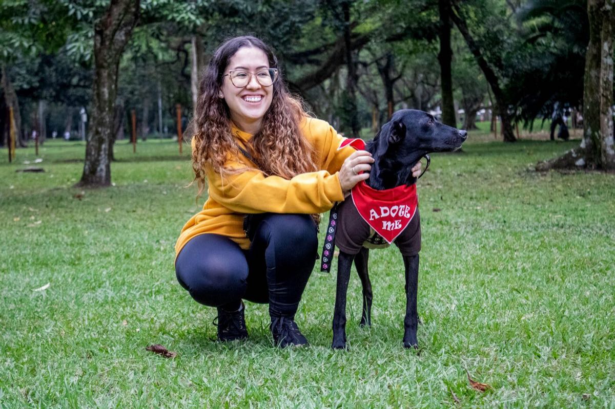 A woman kneeling in a park beside a black dog wearing an adoption bandana.
